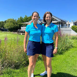 Image of two female golfers wearing the Ocean Meets Green TIDE skirt in navy and the MOANA poloshirt in light blue