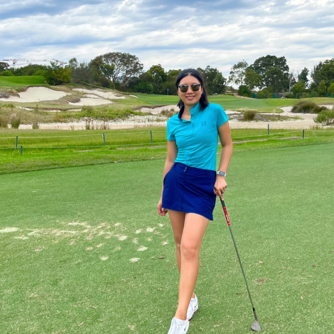 Image of woman wearing Ocean Meets Green Moana golf polo in green and Tide skirt in navy, standing on a teebox