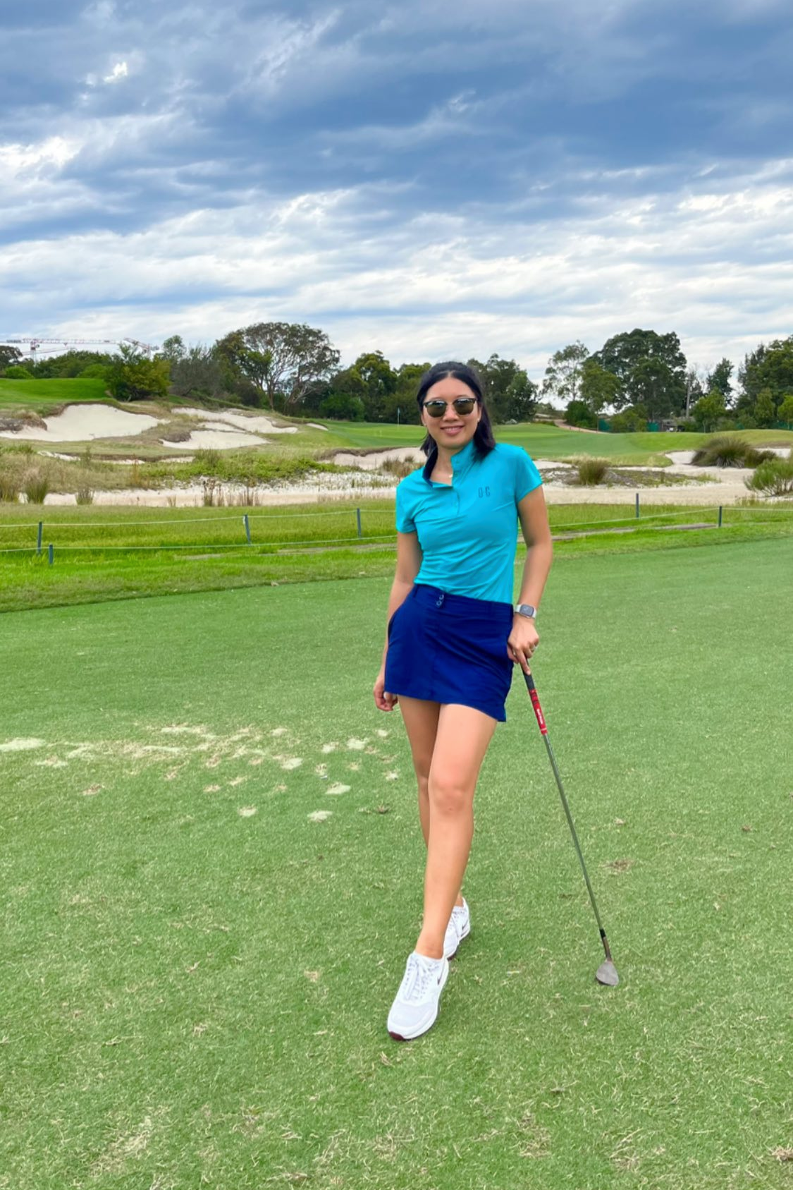 Image of woman wearing Ocean Meets Green Moana golf polo in green and Tide skirt in navy, standing on a teebox