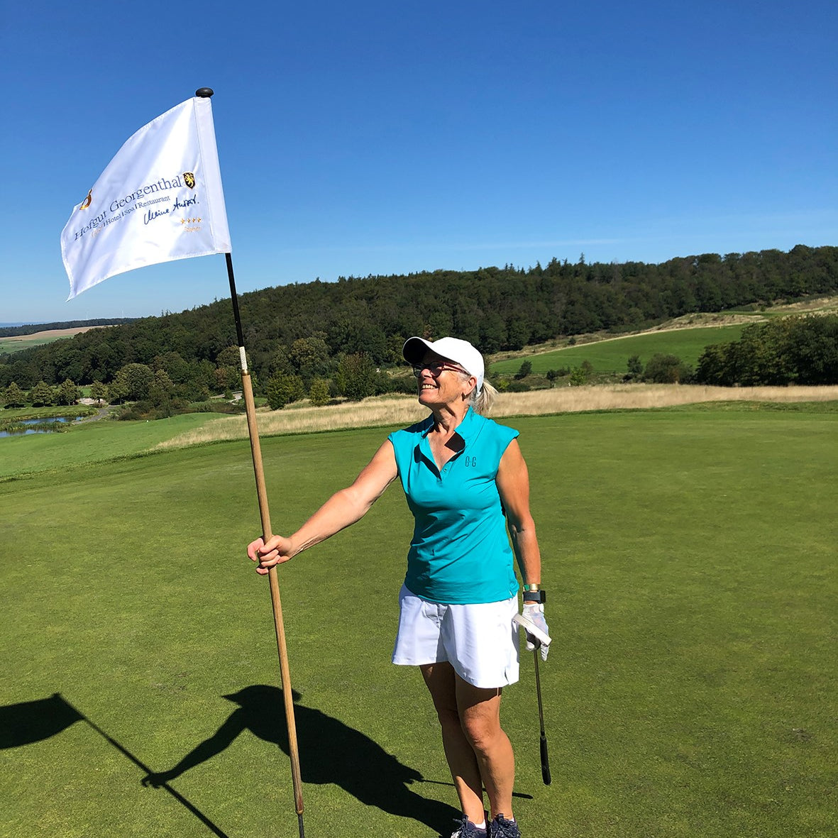 Image of woman wearing Ocean Meets Green Moana golf polo in green and Tide skirt in white, standing on a putting green