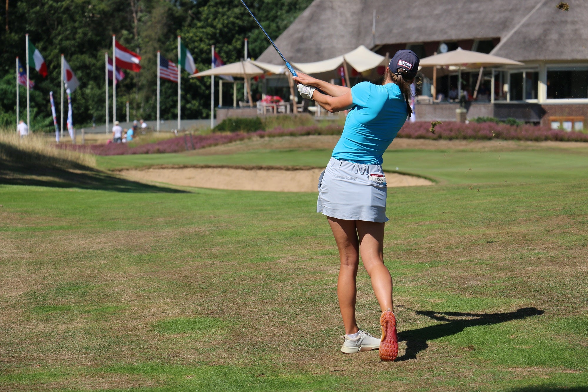 Image of woman wearing Ocean Meets Green Moana golf polo in light blue and Tide skirt in grey, hitting an approach shot