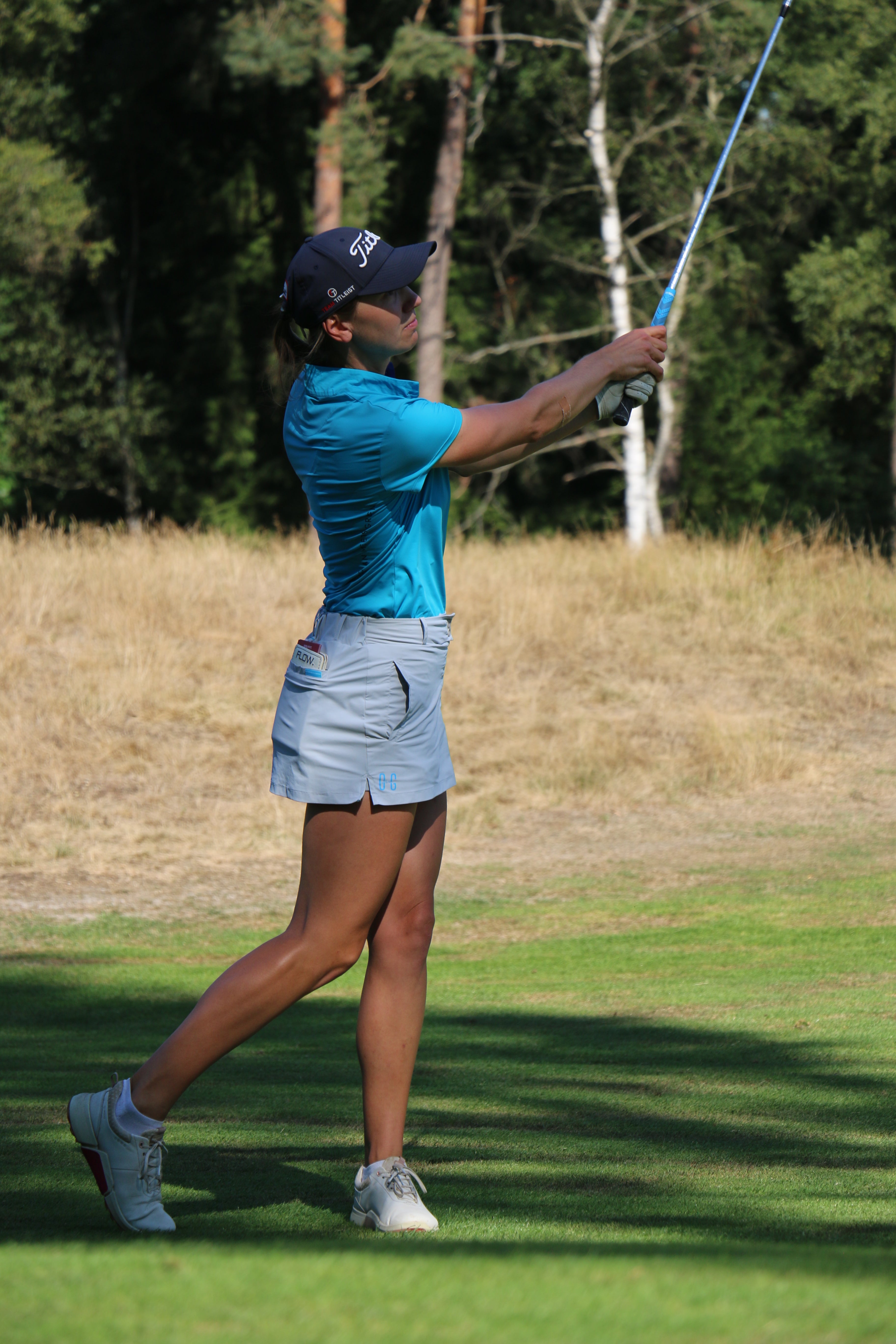 Image of woman wearing Ocean Meets Green Moana golf polo in light blue and Tide skirt in grey, hitting an iron shot